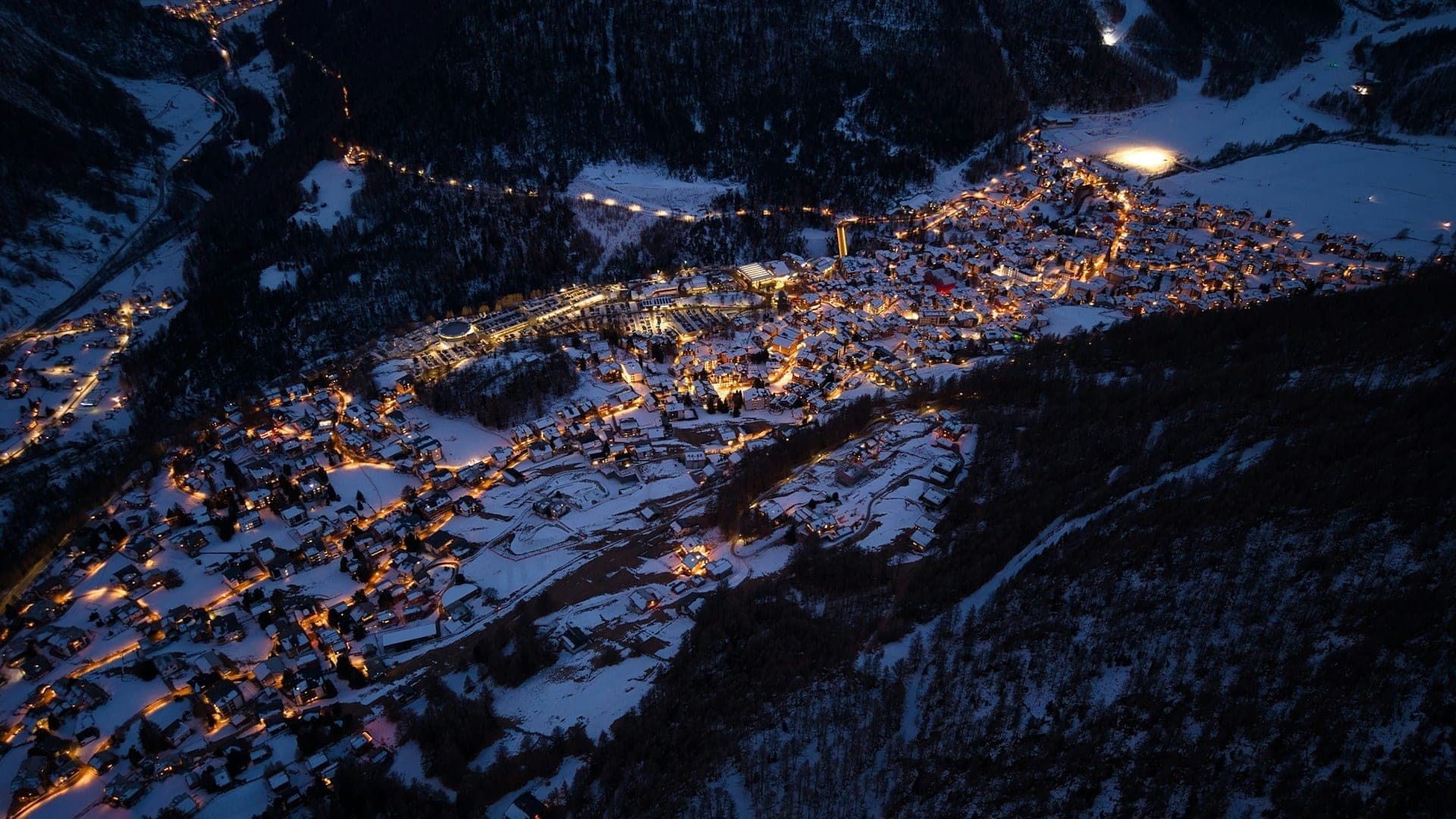 Snow-covered resort village at dusk with lit windows and a chairlift line above town
